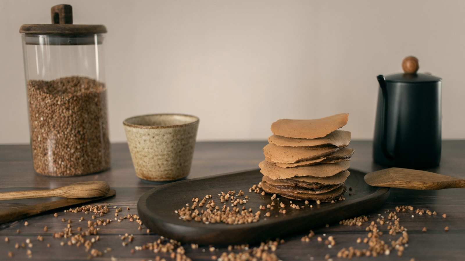 Un barattolo di grano saraceno e una pila di crêpe di grano saraceno su un vassoio di legno, con chicchi sparsi