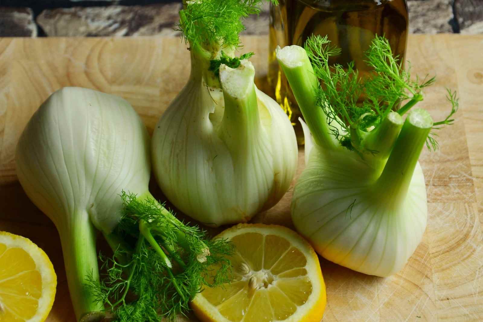 Finocchi freschi con le loro foglie e mezzi limoni su un tagliere di legno, con una bottiglia d'olio dietro