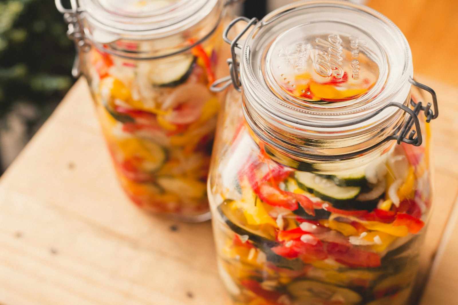 Due vasetti di vetro pieni di peperoni, zucchine e cipolle in conserva, su un piano di legno