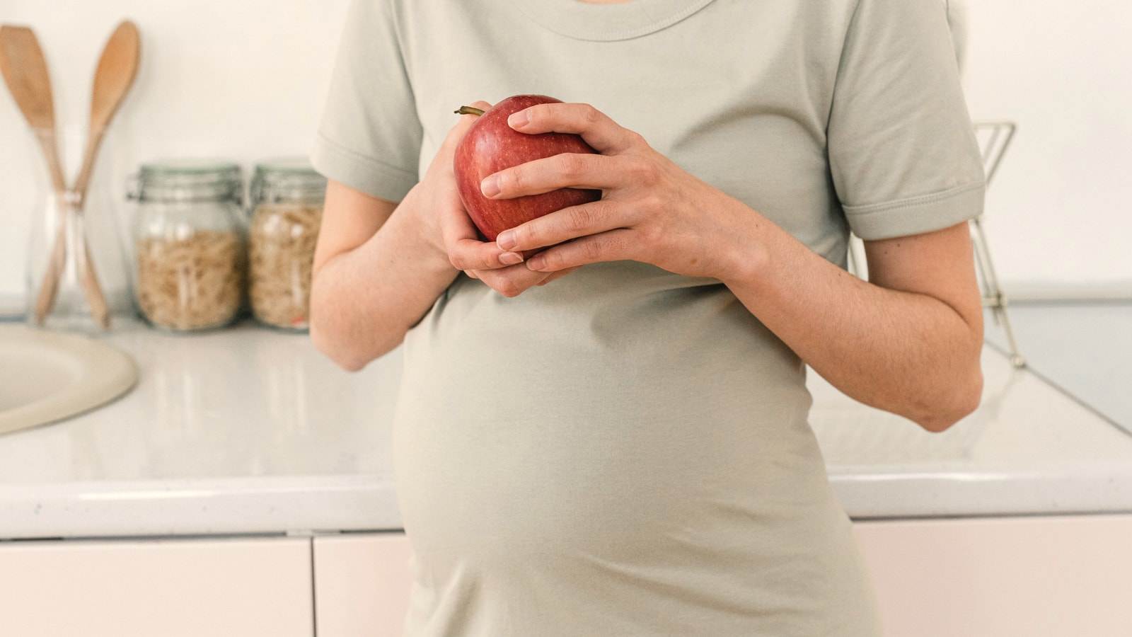 Una donna incinta tiene una mela rossa davanti al ventre, in cucina
