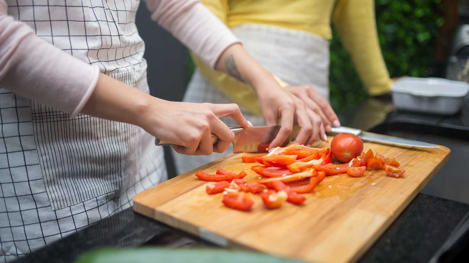 Due persone in grembiule tagliano peperoni rossi e pomodori su un tagliere di legno