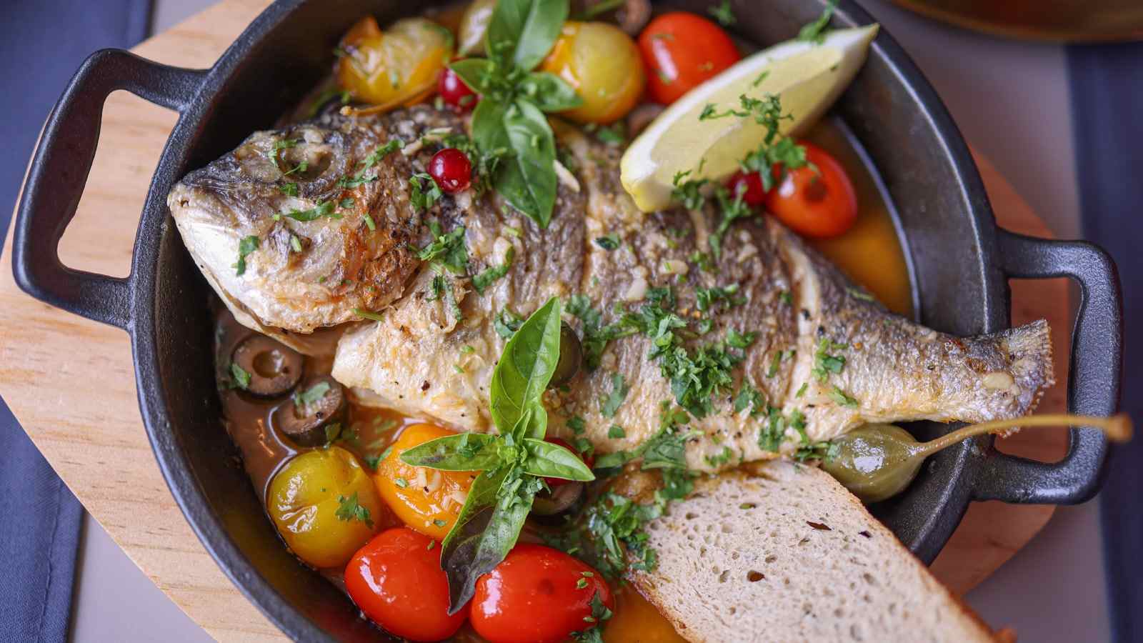 Un pesce al forno con pomodorini, basilico, olive e limone in una pentola di ghisa, con una fetta di pane