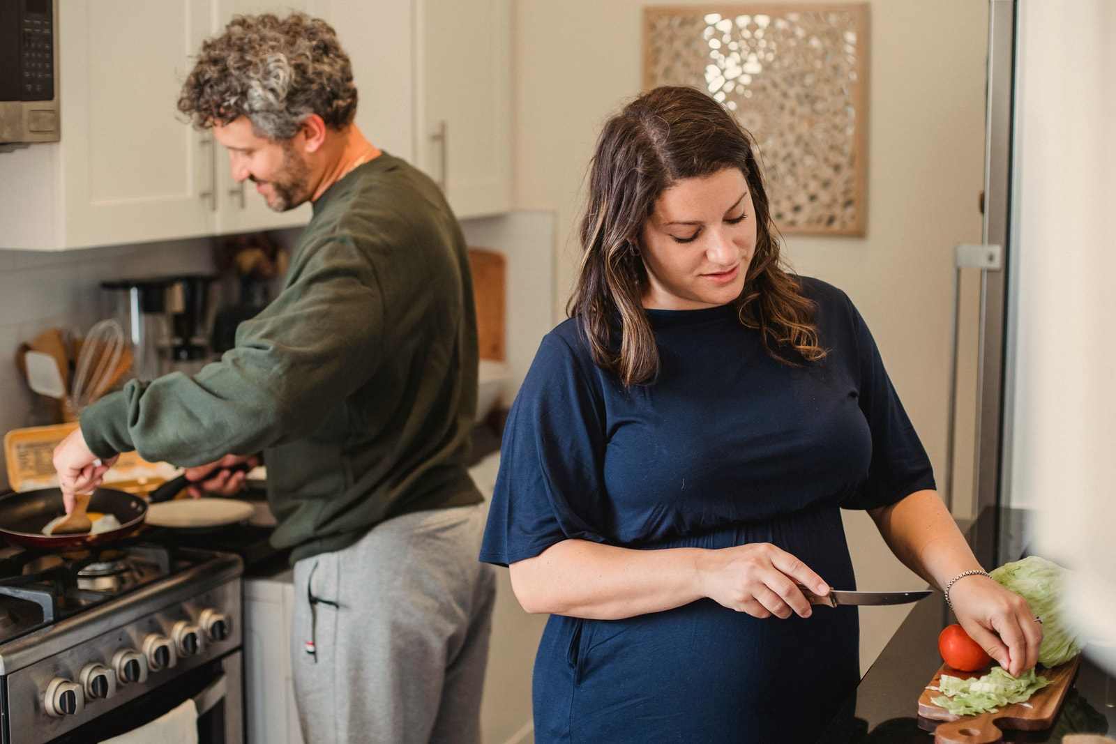 Una donna incinta taglia le verdure su un tagliere mentre il compagno cucina ai fornelli