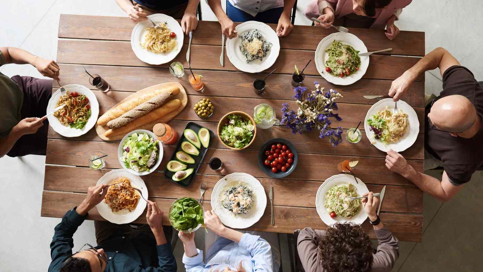Una tavola di legno vista dall'alto, con pasta, insalate, pane e le persone sedute attorno
